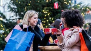 two ladies clinking mugs of hot cocoa while holding shopping bags with blurred christmas tree in background